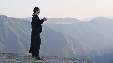 Güneşin doğuşunda bir dağda geleneksel siyah bir kıyafetle Tai Chi uygulayan bir kadın, kollarını meditasyon pozuyla açmış, denge, enerji ve doğadaki farkındalığa odaklanıyor. Yavaş çekim