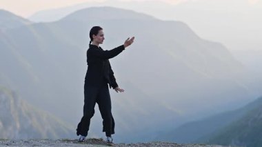 Güneşin doğuşunda bir dağda geleneksel siyah bir kıyafetle Tai Chi uygulayan bir kadın, kollarını meditasyon pozuyla açmış, denge, enerji ve doğadaki farkındalığa odaklanıyor. Yavaş çekim