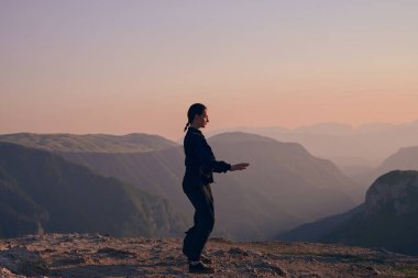 Güneşin doğuşunda bir dağda geleneksel siyah bir kıyafetle Tai Chi çalışması yapan bir kadın, kollarını meditasyon pozunda uzatarak, doğada denge, enerji ve farkındalık üzerine odaklanır.. 