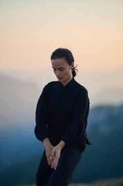 Güneşin doğuşunda bir dağda geleneksel siyah bir kıyafetle Tai Chi çalışması yapan bir kadın, kollarını meditasyon pozunda uzatarak, doğada denge, enerji ve farkındalık üzerine odaklanır.. 