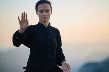 Güneşin doğuşunda bir dağda geleneksel siyah bir kıyafetle Tai Chi çalışması yapan bir kadın, kollarını meditasyon pozunda uzatarak, doğada denge, enerji ve farkındalık üzerine odaklanır.. 