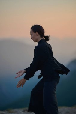 Güneşin doğuşunda bir dağda geleneksel siyah bir kıyafetle Tai Chi çalışması yapan bir kadın, kollarını meditasyon pozunda uzatarak, doğada denge, enerji ve farkındalık üzerine odaklanır.. 
