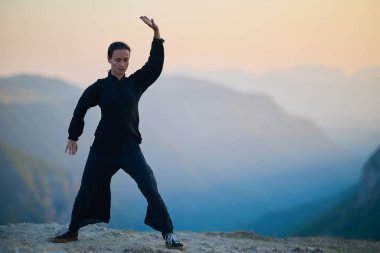 Güneşin doğuşunda bir dağda geleneksel siyah bir kıyafetle Tai Chi çalışması yapan bir kadın, kollarını meditasyon pozunda uzatarak, doğada denge, enerji ve farkındalık üzerine odaklanır.. 