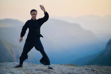 Güneşin doğuşunda bir dağda geleneksel siyah bir kıyafetle Tai Chi çalışması yapan bir kadın, kollarını meditasyon pozunda uzatarak, doğada denge, enerji ve farkındalık üzerine odaklanır.. 