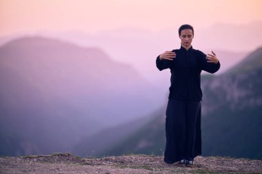 Güneşin doğuşunda bir dağda geleneksel siyah bir kıyafetle Tai Chi çalışması yapan bir kadın, kollarını meditasyon pozunda uzatarak, doğada denge, enerji ve farkındalık üzerine odaklanır.. 