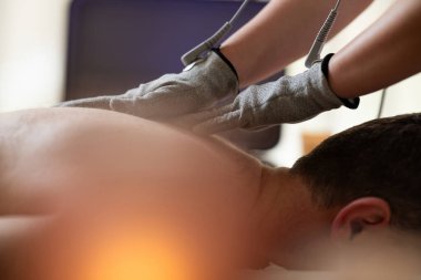 Fizyoterapi seansı sırasında bir adama özel iletken eldivenlerle elektroterapi uygulayan bir terapiste yakından bakın. Modern rehabilitasyon kavramı, ağrı kesici, kas stimülasyonu ve...