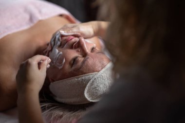 Profesyonel bir terapist yüz temizleme ameliyatı sırasında köpük uygularken yetişkin bir kadının havlu ve saç bandıyla tedavi yatağına uzanması. Cilt bakımı ve sağlık konsepti.