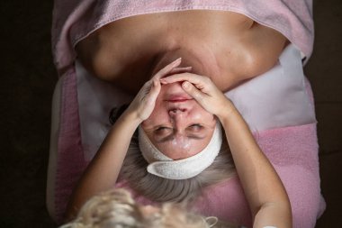 Profesyonel bir terapist yüz temizleme ameliyatı sırasında köpük uygularken yetişkin bir kadının havlu ve saç bandıyla tedavi yatağına uzanması. Cilt bakımı ve sağlık konsepti.