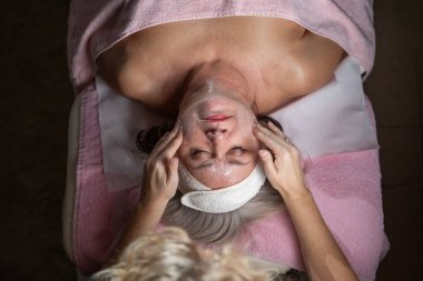 Profesyonel bir terapist yüz temizleme ameliyatı sırasında köpük uygularken yetişkin bir kadının havlu ve saç bandıyla tedavi yatağına uzanması. Cilt bakımı ve sağlık konsepti.