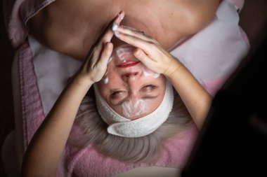 Profesyonel bir terapist yüz temizleme ameliyatı sırasında köpük uygularken yetişkin bir kadının havlu ve saç bandıyla tedavi yatağına uzanması. Cilt bakımı ve sağlık konsepti.