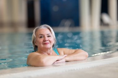 Yüzme havuzunda havuz kenarında dinlenen gülümseyen, son sınıf öğrencisi bir kadın. Aktif yaşam tarzı, sağlık, hidroterapi ve yaşlı yetişkinler için boş zaman kavramı