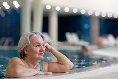 Yüzme havuzunda havuz kenarında dinlenen gülümseyen, son sınıf öğrencisi bir kadın. Aktif yaşam tarzı, sağlık, hidroterapi ve yaşlı yetişkinler için boş zaman kavramı
