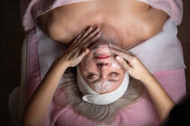 Profesyonel bir terapist yüz temizleme ameliyatı sırasında köpük uygularken yetişkin bir kadının havlu ve saç bandıyla tedavi yatağına uzanması. Cilt bakımı ve sağlık konsepti.