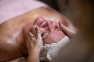 Terapist yüzüne nazikçe masaj yaparken yetişkin bir kadının saç bandıyla tedavi yatağına uzanması. Spa güzelliği ve sağlık konsepti rahatlama, cilt bakımı, gençleşme ve