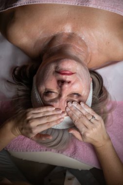 Profesyonel bir terapist yüz temizleme ameliyatı sırasında köpük uygularken yetişkin bir kadının havlu ve saç bandıyla tedavi yatağına uzanması. Cilt bakımı ve sağlık konsepti.