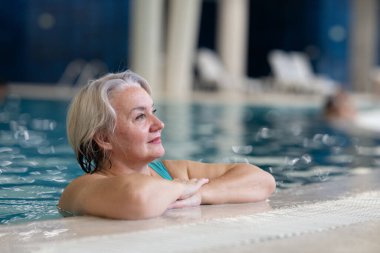 Yüzme havuzunda havuz kenarında dinlenen gülümseyen, son sınıf öğrencisi bir kadın. Aktif yaşam tarzı, sağlık, hidroterapi ve yaşlı yetişkinler için boş zaman kavramı