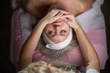 Profesyonel bir terapist yüz temizleme ameliyatı sırasında köpük uygularken yetişkin bir kadının havlu ve saç bandıyla tedavi yatağına uzanması. Cilt bakımı ve sağlık konsepti.