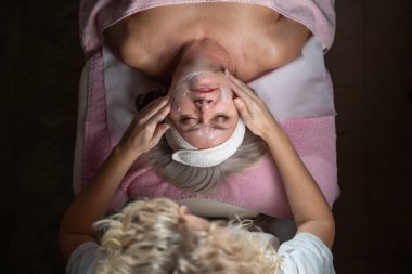 Profesyonel bir terapist yüz temizleme ameliyatı sırasında köpük uygularken yetişkin bir kadının havlu ve saç bandıyla tedavi yatağına uzanması. Cilt bakımı ve sağlık konsepti.