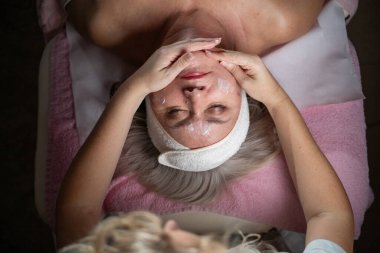 Profesyonel bir terapist yüz temizleme ameliyatı sırasında köpük uygularken yetişkin bir kadının havlu ve saç bandıyla tedavi yatağına uzanması. Cilt bakımı ve sağlık konsepti.