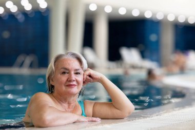 Yüzme havuzunda havuz kenarında dinlenen gülümseyen, son sınıf öğrencisi bir kadın. Aktif yaşam tarzı, sağlık, hidroterapi ve yaşlı yetişkinler için boş zaman kavramı