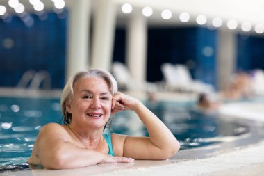 Yüzme havuzunda havuz kenarında dinlenen gülümseyen, son sınıf öğrencisi bir kadın. Aktif yaşam tarzı, sağlık, hidroterapi ve yaşlı yetişkinler için boş zaman kavramı