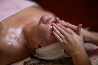 Terapist yüzüne nazikçe masaj yaparken yetişkin bir kadının saç bandıyla tedavi yatağına uzanması. Spa güzelliği ve sağlık konsepti rahatlama, cilt bakımı, gençleşme ve