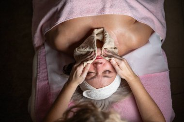 Profesyonel bir terapist yüz temizleme ameliyatı sırasında köpük uygularken yetişkin bir kadının havlu ve saç bandıyla tedavi yatağına uzanması. Cilt bakımı ve sağlık konsepti.