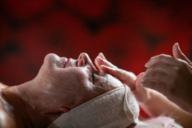 Profesyonel bir terapist yüz temizleme ameliyatı sırasında köpük uygularken yetişkin bir kadının havlu ve saç bandıyla tedavi yatağına uzanması. Cilt bakımı ve sağlık konsepti.