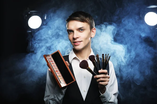 Professional makeup artist posing in studio, holds a palette of shadows and brush.Against the background of smoke. — Stock Photo, Image