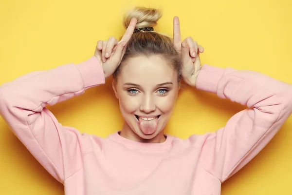 Portrait of playful young pretty blond woman showing horns and making faces. Isolated on yellow background. — Stock Photo, Image