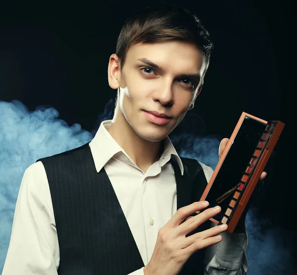 Male make-up artist staying on dark background, holding professional brush and shadows palette, looking at camera — Stock Photo, Image
