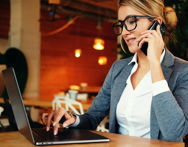 Vidám fiatal gyönyörű nő szemüvegben beszél a mobiltelefon és a laptop mosollyal, miközben ül a munkahelyén. — Stock Fotó