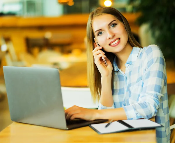 Jeune belle fille blonde assise dans un café et travaillant sur ordinateur. Image d'une femme heureuse utilisant un ordinateur portable et un smartphone dans un café . — Photo