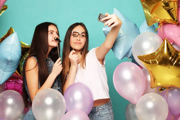 Två tonårstjejer med färgglada ballonger gör selfie på en telefon. Blå bakgrund. — Stockfoto