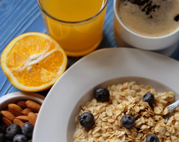 Mingau de aveia com mirtilos frescos, amêndoas, café e suco de laranja em tigela branca na mesa azul. Café da manhã saudável, alimentação saudável, conceito de comida vegan . — Fotografia de Stock