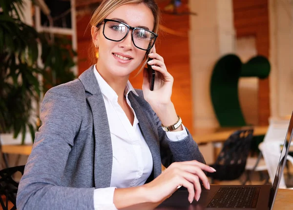 Buena charla de negocios. joven hermosa mujer en gafas hablando en el teléfono móvil y el uso de la computadora portátil con sonrisa mientras se sienta en su lugar de trabajo . — Foto de Stock