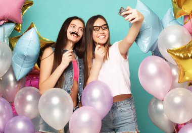 Renkli balonları olan iki genç kız arkadaş telefonda selfie çekiyorlar. Mavi arkaplan.
