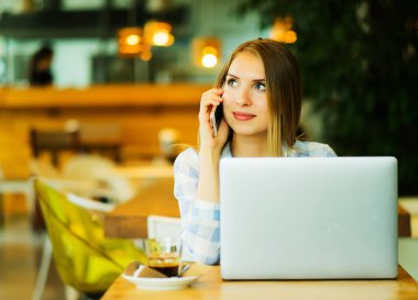 Bir kafede otururken ve bilgisayarda çalışan genç güzel sarışın kız. Kafede dizüstü bilgisayar ve akıllı telefon kullanan mutlu kadın görüntüsü. Yaşam tarzı kavramı.