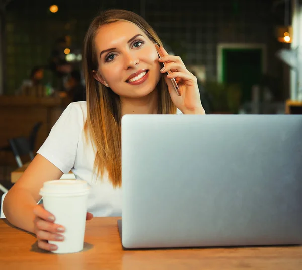 Gyönyörű szőke kislány ül egy kávézóban, és dolgozik a számítógépen. Használ hordozható és smartphone kávézóban boldog nő képe. Életmód fogalom. — Stock Fotó