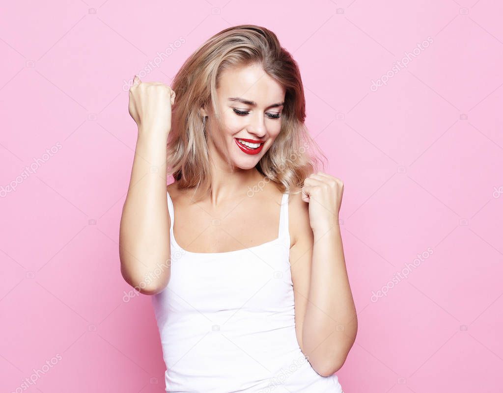 Happy successful young blond woman with raised hands shouting and celebrating success over pink background
