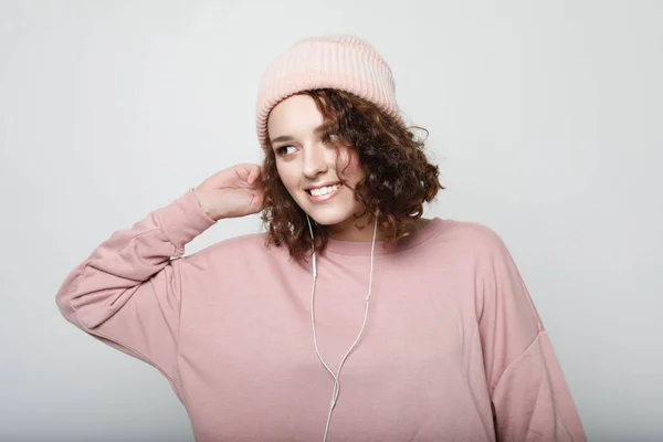 Retrato de una hermosa chica con auriculares escucha música y sonrisas mientras está de pie sobre un fondo blanco con una sudadera y un sombrero rosa. Concepto de estilo de vida . — Foto de Stock
