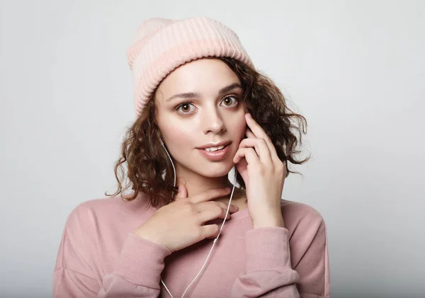 Portret van een mooi meisje in hoofdtelefoon luistert naar muziek en glimlacht terwijl staande op een witte achtergrond in een roze trui en muts. Levensstijl concept. — Stockfoto