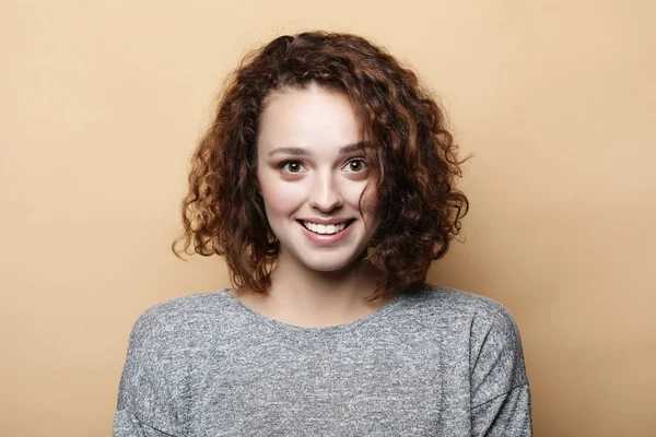 Concepto de personas, felicidad y expresiones faciales. Mujer rizada bastante joven con linda sonrisa posa sola sobre fondo beige . —  Fotos de Stock