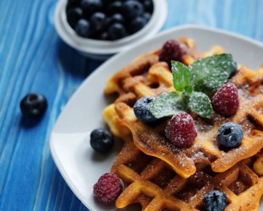 Geleneksel Belçika waffleları taze böğürtlenler ve beyaz tabakta şeker tozu, mavi ahşap arka plan. Düz konum, üst görünüm, kopyalama alanı.