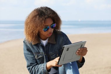 Plajda tableti olan Afrikalı Amerikalı genç bir kadın.