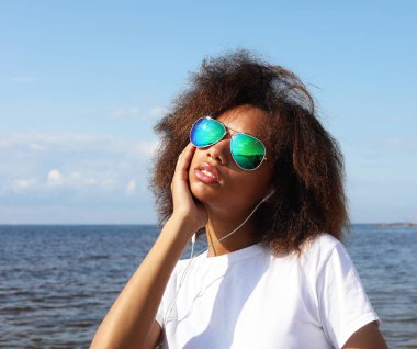 Güneş gözlüklü, kulaklıklı, cep telefonuyla müzik dinleyen büyüleyici afro-amerikalı genç kadın. Sıradan giyin..