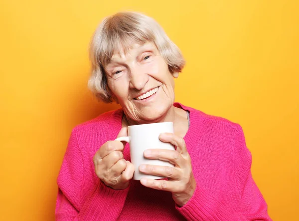 Portrait of old excited lady smiling laughing, holding cup drinking coffee, tea, beverage on yellow background — Stock Photo, Image