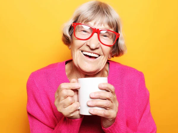 Retrato de velha senhora animada sorrindo rindo, segurando xícara bebendo café, chá, bebida no fundo amarelo — Fotografia de Stock