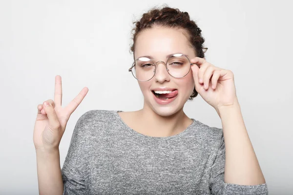 Giovane donna riccia con gli occhiali vestiti con disinvoltura facendo facce alla macchina fotografica, lampeggiando, sporgendo la lingua. Ragazza positiva divertirsi in casa . — Foto Stock