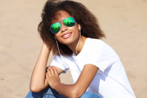 Encantadora jovem afro-americana incrível em óculos de sol ouvindo música em fones de ouvido em seu telefone celular. Vestido casual . — Fotografia de Stock